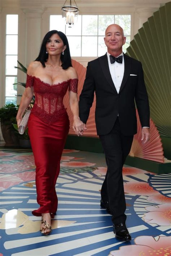Jeff Bezos is wearing a white suit and a black shirt and Mackenzie Scott is wearing a dress with a lot of patterns on it. 